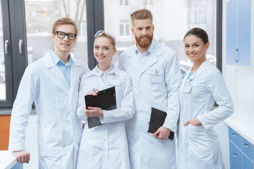 Team of young professional scientists in white coats smiling at camera in laboratory