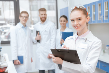 Obraz premium Young smiling woman scientist with clipboard standing near colleagues in lab