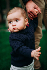 beautiful baby boy staying holding father's hand among green grass on spring lawn