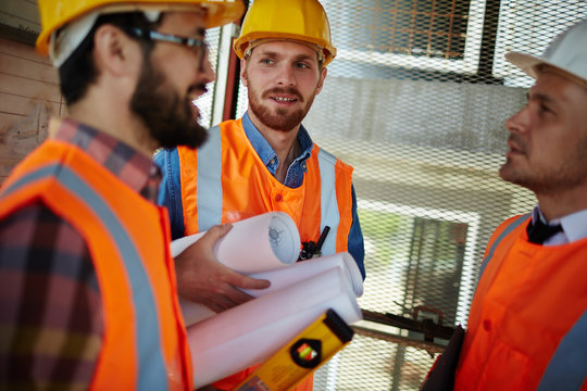 Group of three workmen wearing protective helmets and vests standing in elevator shaft on construction site discussing development progress