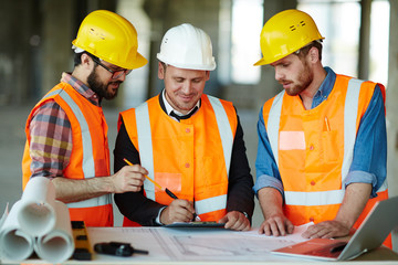 Team of construction workers wearing protective helmets and vests discussing project details with...