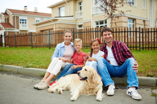 Portrait of happy family with two children, boy and girl, and their golden retriever dog sitting outside on green grass lawn in front of their new big house