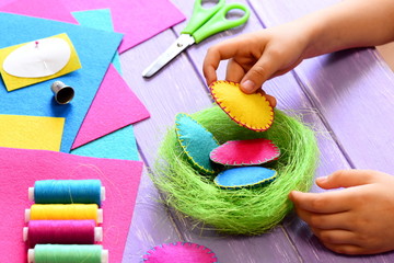 Naklejka premium Small child does Easter decor. Child holds a felt egg in hand. Colorful felt Easter eggs and sisal nest, sewing tools and materials on a table. Simple and creative Easter decor idea