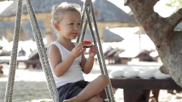 Little cute baby girl sitting on the swing and eat red watermelon at sandy beach