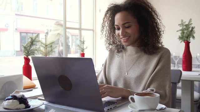 young happy smiling afro american woman with curly hair use laptop for working remotely, taking notes or for chat with friends in social networks while sitting in cafe during sunny day