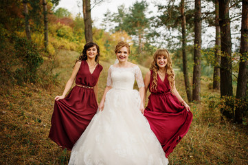 Bride with bridesmaids on red dresses at autumn pine wood.