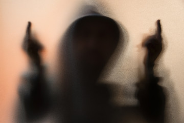 Shadow blur of horror man in jacket with hood.Middle finger symbol fuck you on the glass.Dangerous man behind the frosted glass.Mystery man.Halloween background.Blur picture.