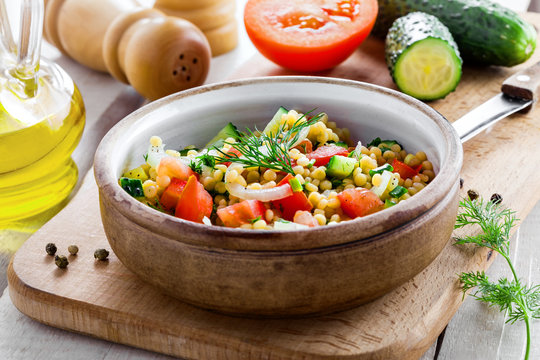 Salad made of couscous with vegetables in a bowl for healthy meal. Cooking of traditional Israeli Ptitim for lunch. Moroccan food and ingredients on a table.