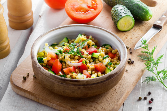 Healthy salad for breakfast made of couscous, tomatoes, cucumber and onion on a table. Cooking of traditional Israeli Ptitim meal and ingredients on a white wooden table. Moroccan cuisine food.