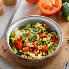 Salad made of couscous with vegetables in a bowl for healthy meal. Cooking of traditional Israeli Ptitim for lunch. Moroccan food and ingredients on a table. Top view.