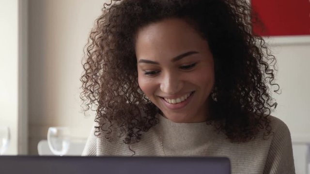 young happy smiling afro american woman with curly hair use laptop for working remotely, taking notes or for chat with friends in social networks while sitting in cafe during sunny day