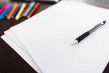 Laptop, white paper, pencil and 12 color markers on the wooden working desk in soft focus, focus on pen.