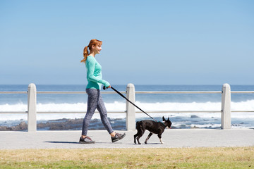fit woman smiling and walking dog on path by sea