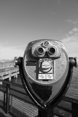 Tower viewer binoculars on Liberty Island looking towards Manhattan, New York.  Pier and Manhattan skyline in the distance. Black and white, focus on tower viewer.