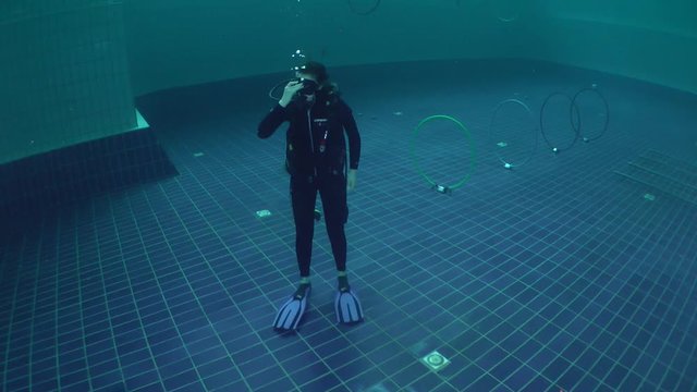 The girl-diver in scuba gear standing on the bottom of the pool, wide shot.
