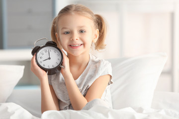 Cute little girl with alarm clock sitting on bed
