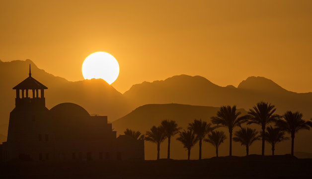The building in the oriental style with a tower and silhouettes of palm trees at sunset in the mountainous desert.