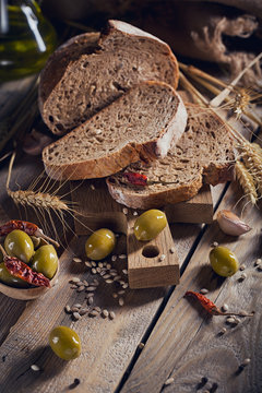 Fresh multigrain crusty bread, green olives, chilly pepper and wheat ears on a rustic wooden table.