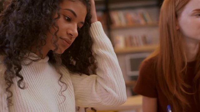 Curly teenage female in white sweater sitting at library with redhead friend making notes in slowmotion