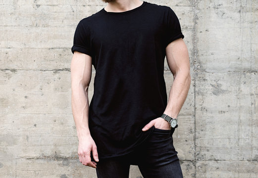 Closeup view of young bearded man wearing black tshirt and jeans posing in center of modern city. Empty concrete wall on the background. Hotizontal mockup.