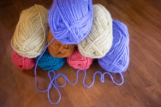 Eight skeins of pink, cream, gold, turquoise and lavender yarn from above on wooden table. Lavender yarn spells the work "yarn" in cursive.