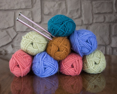 Stack of Eight Skeins of Lavender, Turquoise, Gold, Coral Pink and Pale Green Wool Yarn with Knitting Needles through One Skein On Wooden Table with Stone Wall in Background