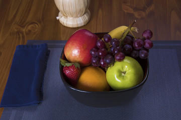 Colorful fruit in a bowl rests on a blue place mat on a dark wood background with a Chianti bottle in the background