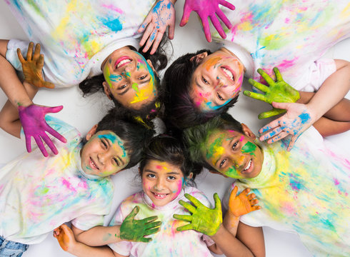 Smiling Indian Kids Celebrating Or Playing Holi Festival With Colourful Body And Faces Making Round Or Holding Sweets And Colour Called Gulal