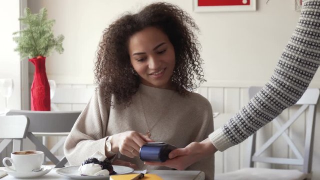 young smiling afro amarican woman with curly hair pay for coffee by card paypass using electronic payment terminal in cafe and drink her coffee