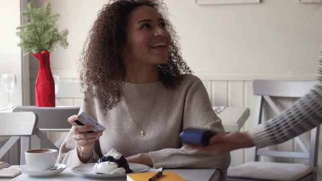 young smiling afro amarican woman with curly hair pay for coffee by card paypass using electronic payment terminal in cafe and drink her coffee