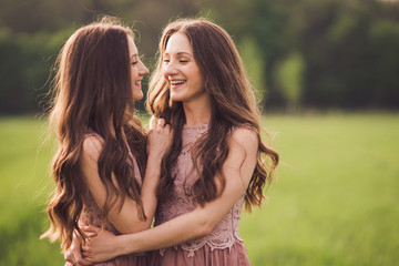 Beautiful happy young twins sisters in long evening dresses hugging in green spring field in sunshine. Having fun together, positive emotions, bright colors. Copy space.