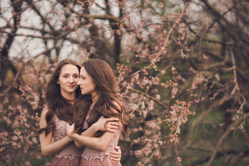 Beautiful happy young twins sisters in long evening dresses enjoying smell in a blooming spring garden. Having fun together, positive emotions, bright colors. Copy space.
