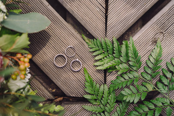 Botanic bridal chic. Bouquet, wedding rings and engagement ring on wooden background with greenery and fern leaves. Copy space, top view, flat lay. Rustic composition.