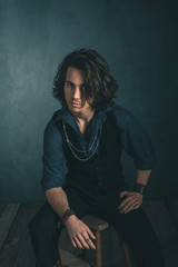 Vintage alternative artistic fashion man. Sitting on wooden stool in studio.