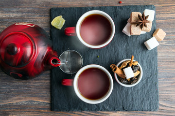 Sooty teapot with strainer, red tea cups, spices and white and brown sugar cubes on slate. Top view.