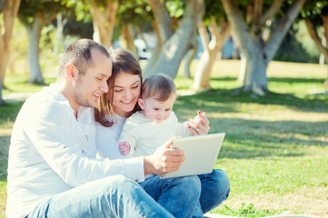 Young parents and little kid using table computer while sitting on grass