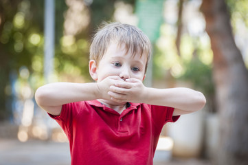 Serious little Caucasian boy closing his mouth with hands. Illustrative image for childhood trauma,...