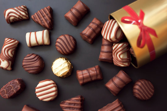 Cioccolatini in diverse forme e colori versato dalla scatola rotonda d'oro con un fiocco rosso su sfondo nero. Tra tutti i dolci hanno un oro avvolto. 