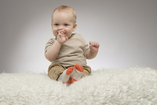 Funny little baby boy with fingers in the mouth, sitting on the white blanket, studio shot, isolated on grey background