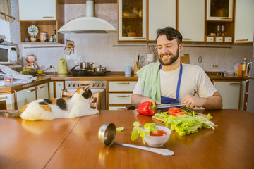 a man cooks in the kitchen beside him sits a funny beautiful cat
