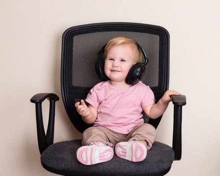 Happy baby in office chair in headphones. Toddler girl listening to music.