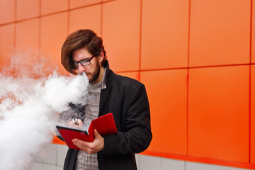 Young hipster man with a beard and glasses smokes an electronic cigarette. He holds a tablet pc. Youth fashion. Delicious pairs. Man makes startup. Business on the Internet.