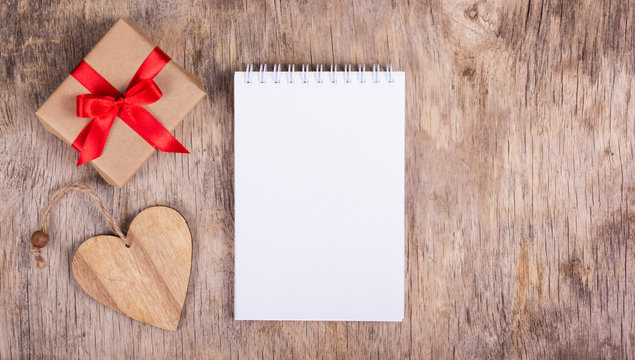 Notepad with blank page, a wooden heart and small gift box with a red bow. Copy space.