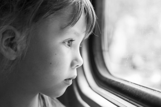 Monochrome portrait of a beautiful girl who looks in the window of the train. Close-up of a sad child looking through window. Black and white photography