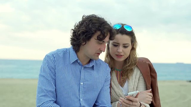 Couple sitting on the beach and browsing internet on smartphone, steadycam shot
