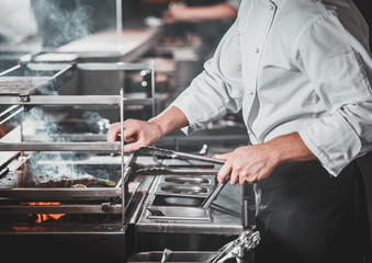White chef in apron standing near the brazier whith coals, only hands. Man cooking beef steak in the interior of modern professional kitchen