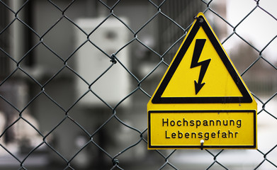 Schwarz-gelbes Warnschild an Maschendrahtzaun - Symbol für Lebensgefahr durch Stromschlag.