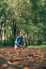 Fit male jogger ties shoes while day training for cross country forest trail race in a nature park.