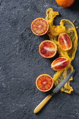 Sliced and whole ripe juicy Sicilian Blood oranges fruits with knife and yellow textile on black concrete texture background. Top view with space