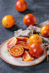 Sliced and whole ripe juicy Sicilian Blood oranges fruits with sackcloth rag on ceramic plate over black concrete texture background.